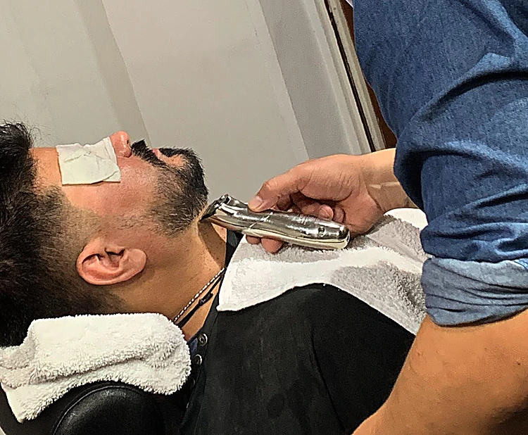 Barbero afeitando la barba de un cliente recostado con toalla blanca al cuello.