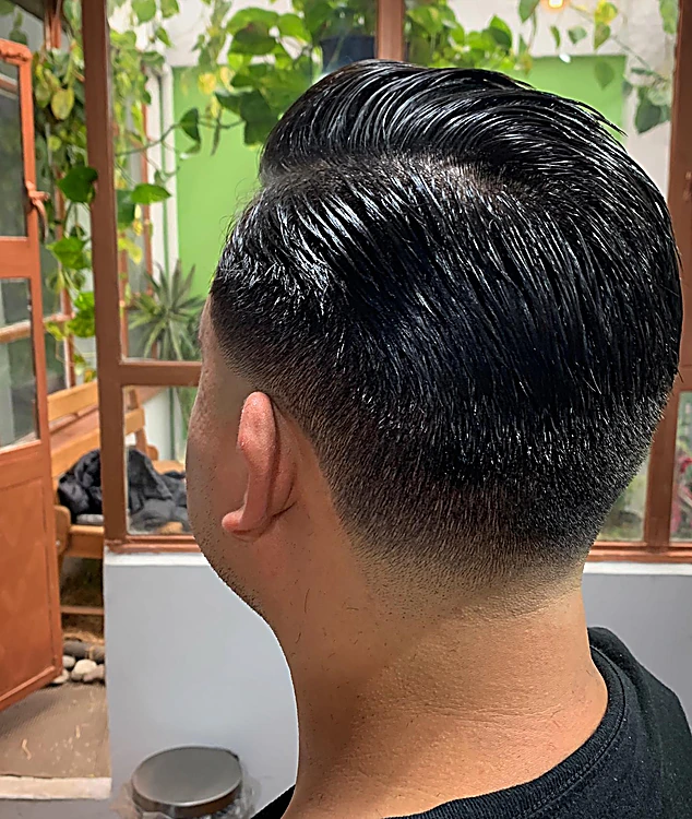 Corte con degradado en la nuca, peinado hacia atrás, con el ventanal y las plantas del patio de fondo.
